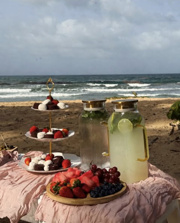 A beach picnic table with chocolate-dipped strawberries, fresh fruit and glass jars of lemonade on blush linen.
